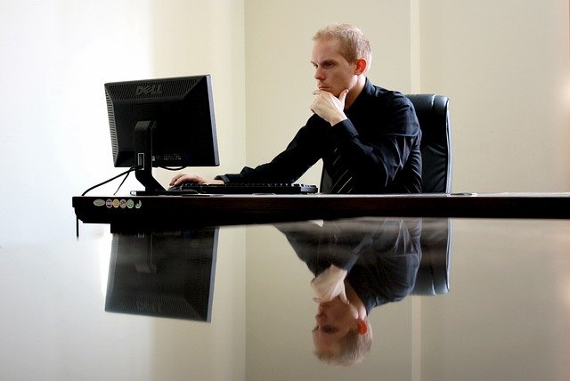 Muž v čiernom roláku sedí pri čiernom stole a pozerá do počítača