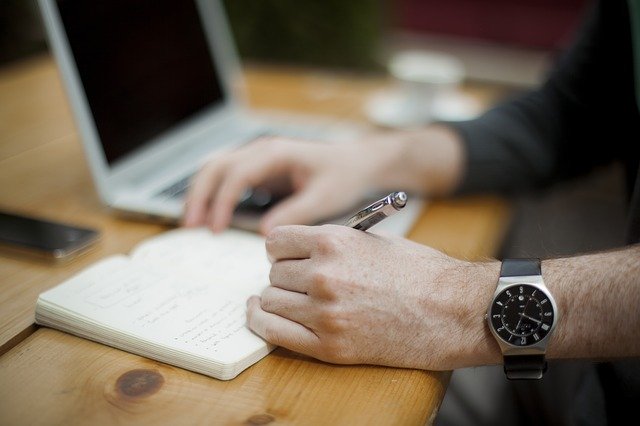 Muž s hodinkami na ruke má na stole notebook a píše do notesa
